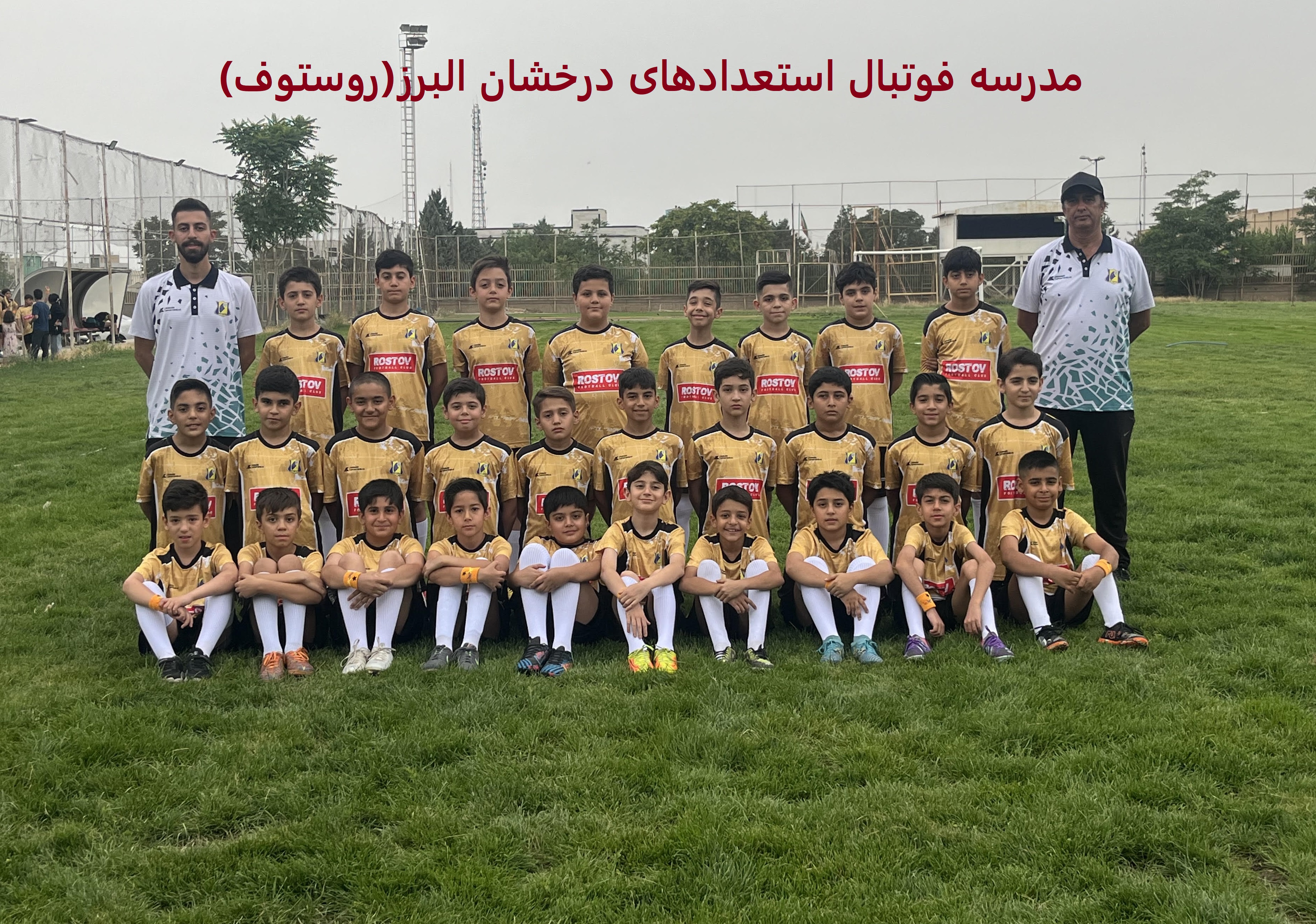 عکس گروهی تیم فوتبال کودکان با لباس‌های زرد و مشکی در زمین چمن، همراه با مربیان ایستاده و بازیکنان نشسته، با پس‌زمینه‌ای از نرده‌های ورزشگاه و درختان