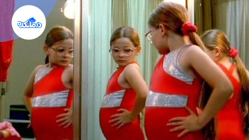 دختر بچه‌ایی که عینک زده و لباس قرمز باشگاه ورزشی را پوشیده است. او جلوی آینه ایستاده و به اندام خود نگاه می‌کند.