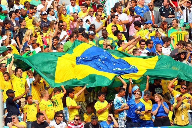 جمعی از طرفداران برزیل در استادیوم فوتبال با در دست داشتن پرچم برزیل شادی می کنند